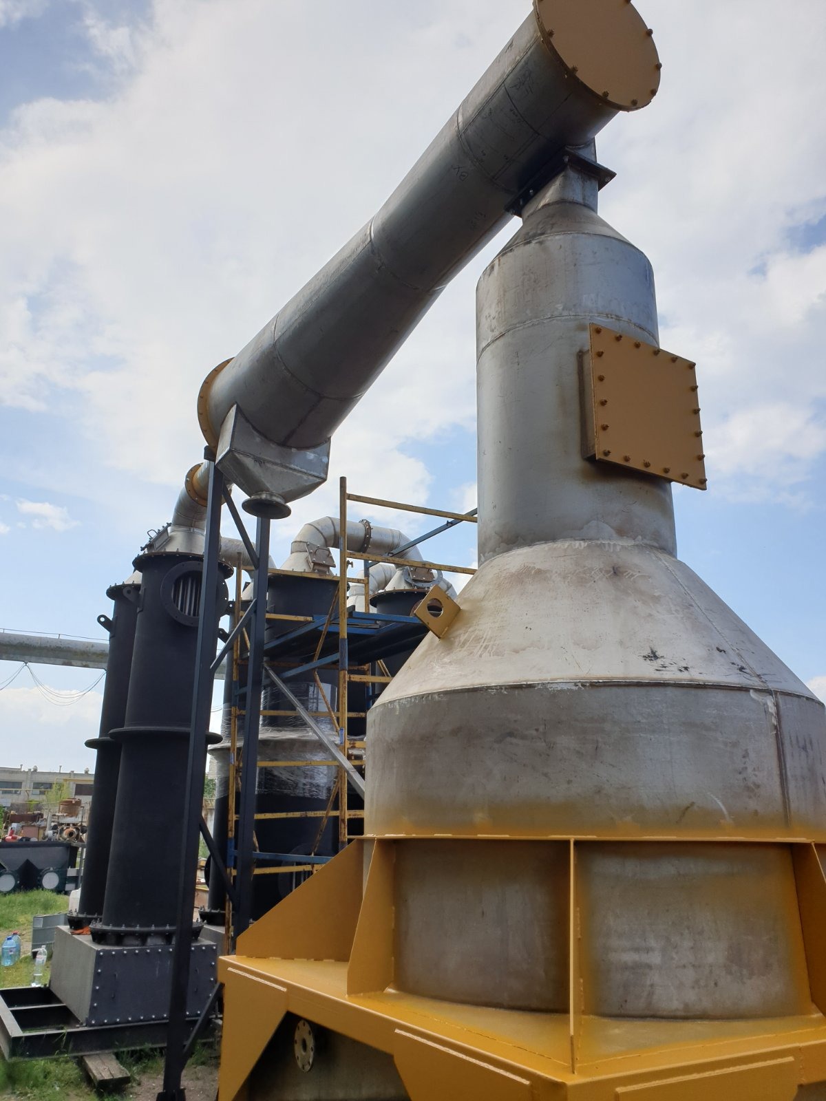 Kompleksowa instalacja przemysłowa z metalowymi rurami, zbiornikami i konstrukcją wsporczą, częściowo pomalowana na żółto, na tle błękitnego nieba.