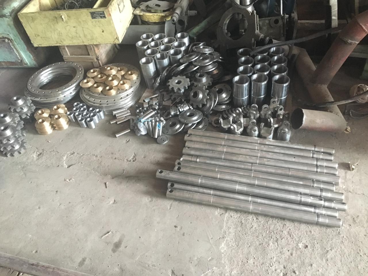 Stalowe i mosiężne elementy po obróbce: pierścienie, tuleje, zębatki i pręty, rozłożone na betonowej posadzce w warsztacie.