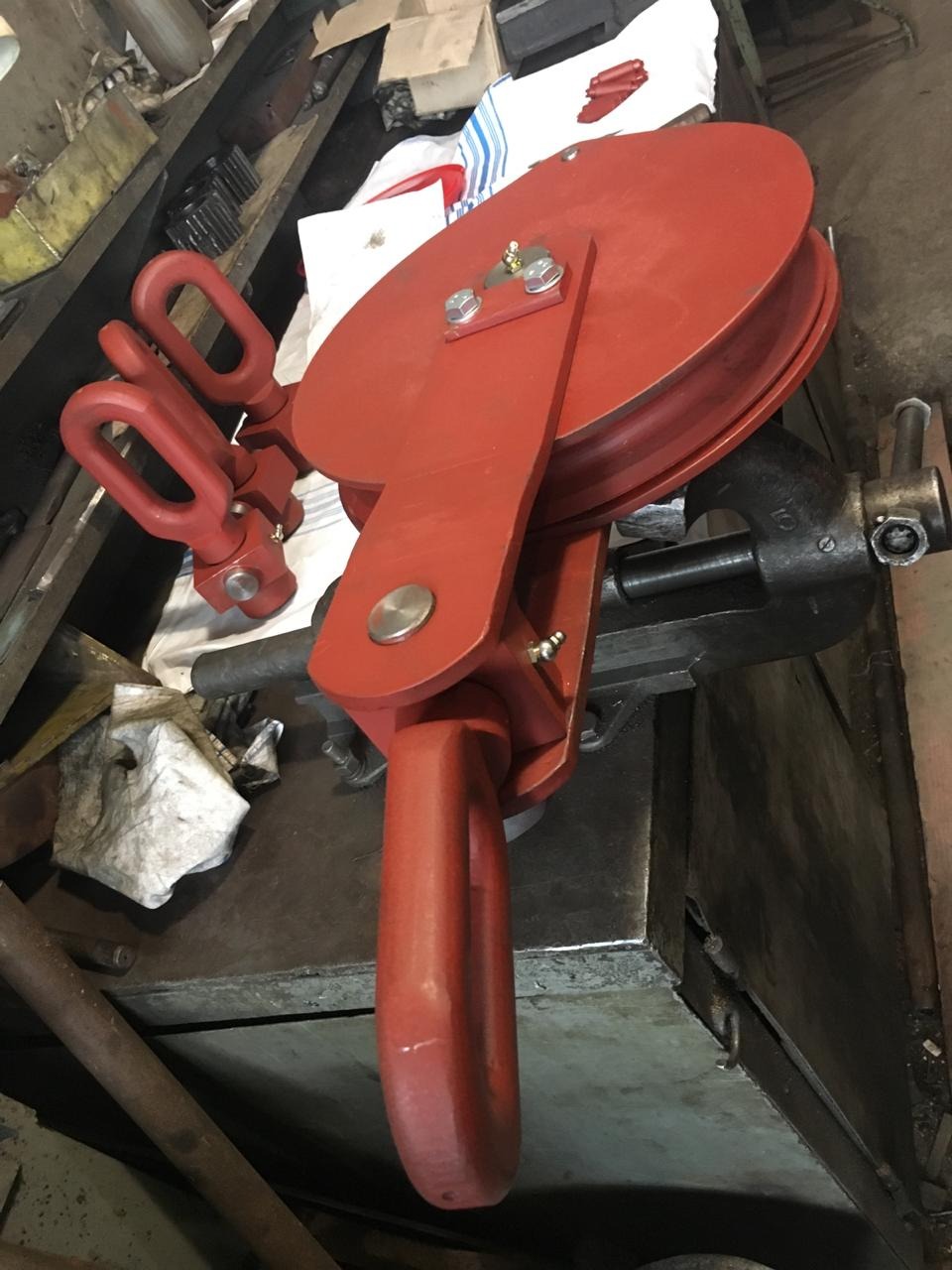 Czerwony bloczek linowy z trzema uchwytami, zamocowany w imadle na warsztatowym stole. Widoczne ślady użytkowania i warsztatowy nieład.