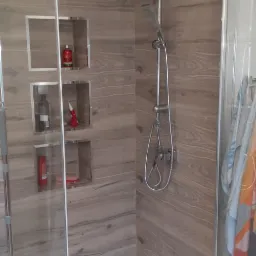 Nowoczesna kabina prysznicowa z panelami imitującymi drewno, wnęki z kosmetykami i akcesoriami, widoczny mop w wiadrze.