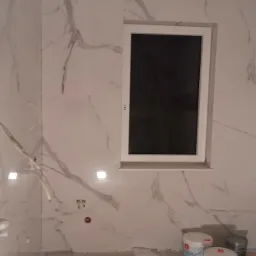 Ściany wyłożone płytkami imitującymi marmur w łazience w trakcie prac wykończeniowych, widoczne puszki z farbą i narzędzia na podłodze.