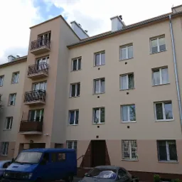 Wielopiętrowy budynek mieszkalny po renowacji elewacji w odcieniach beżu i brązu, z balkonami z metalowymi balustradami, widocznymi zaparkowanymi samochodami i fragmentem zieleni drzew.