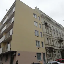 Odnowiona elewacja budynku z balkonami, pomalowana na jasny żółty kolor, sąsiadująca ze starszą kamienicą z dekoracyjnymi detalami architektonicznymi i numerem 22.