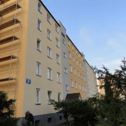 Wielopiętrowy budynek po renowacji elewacji, z fragmentem rusztowania z lewej strony i tabliczką z adresem. Widoczne drzewa iglaste w tle.