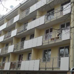 Wielopiętrowy budynek z odnowionymi balkonami z metalowymi balustradami i białymi panelami. Jasna elewacja, widok z dołu, fragment drzewa w kadrze.