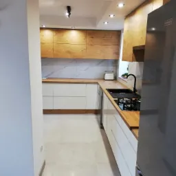 Nowoczesna kuchnia z białymi szafkami dolnymi i drewnopodobnymi górnymi, marmurowy backsplash, czarny zlew i płyta grzewcza, oświetlenie punktowe, widok z korytarza.