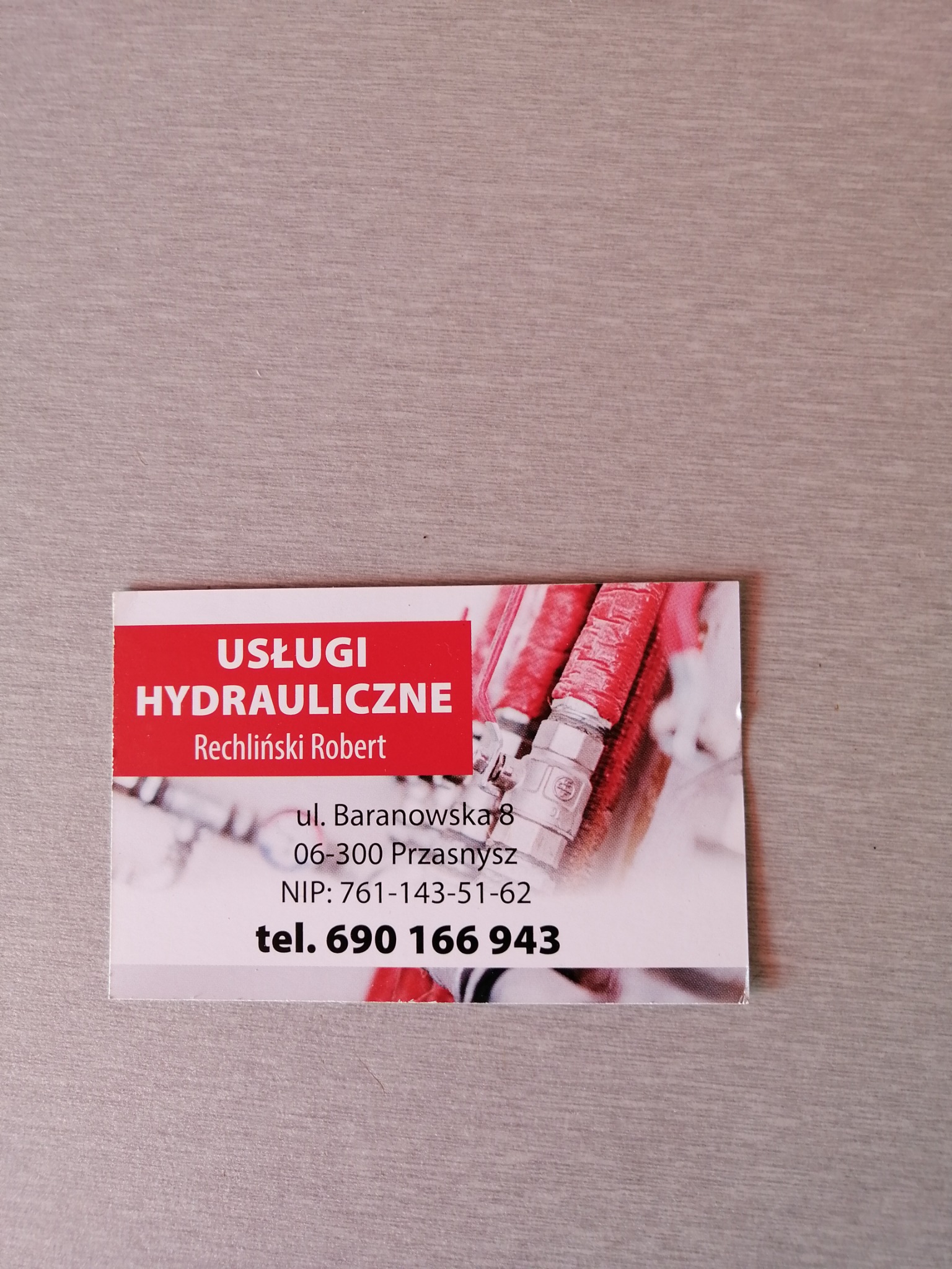Wizytówka firmy hydraulicznej Rechliński Robert z Przasnysza, z adresem ul. Baranowska 8, numerem NIP i telefonem, na tle zdjęcia czerwonych rur hydraulicznych z metalowymi złączkami.