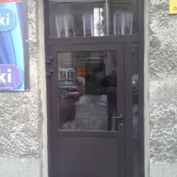 Zewnętrzne, brązowe drzwi wejściowe z szybami, zamontowane w surowej, szarej fasadzie budynku, odbijające ulicę i samochody w szybach.