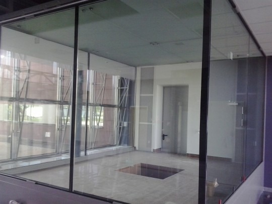 Szklana ścianka działowa z czarnymi profilami, widok na wnętrze biura z oknami i drzwiami.