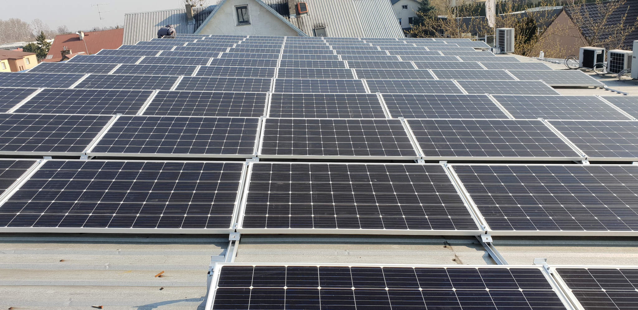 Rozległa instalacja paneli słonecznych na płaskim dachu budynku w Łomży, widoczne moduły fotowoltaiczne z białymi ramami i metalowymi mocowaniami, w tle budynki mieszkalne i urządzenia klimatyzacyjne.