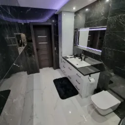 Nowoczesna łazienka z marmurowymi płytkami, białymi meblami łazienkowymi z czarnym blatem, podświetlanym lustrem i podwieszaną toaletą.