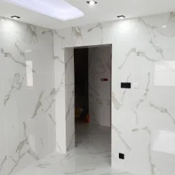 Biała łazienka wykończona płytkami imitującymi marmur, z oświetleniem sufitowym i czarnymi włącznikami, widok na sąsiednie pomieszczenie.