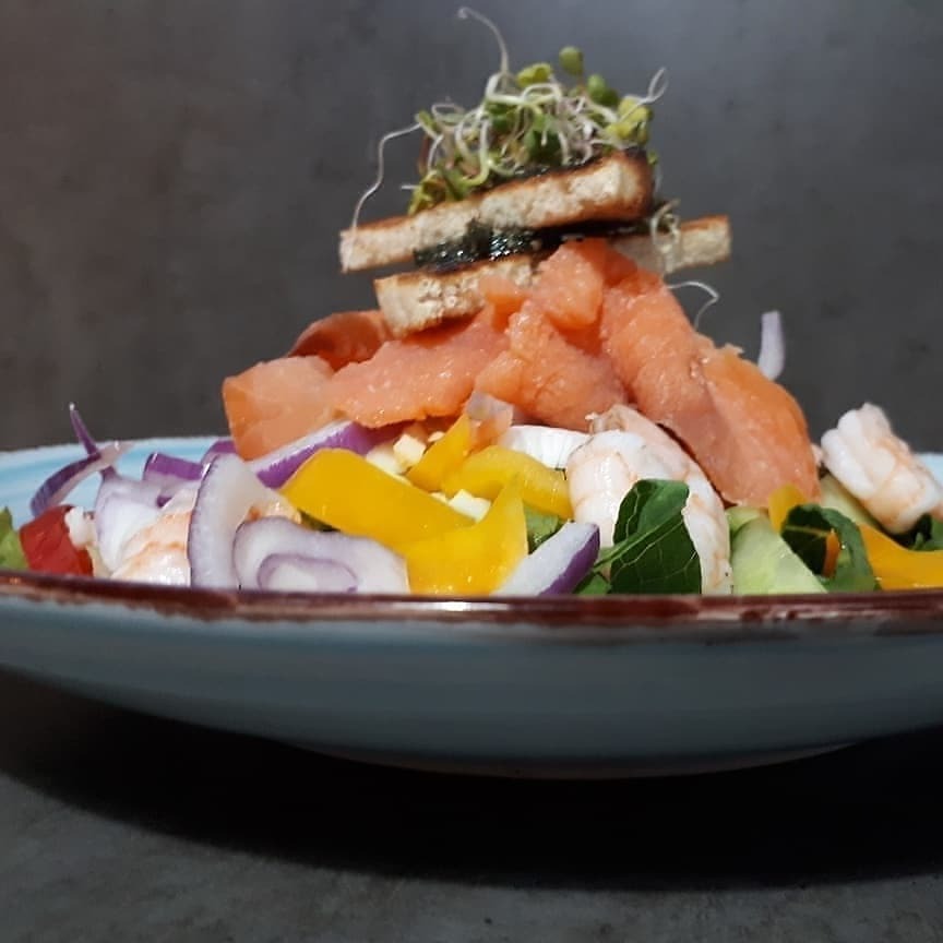 Kolorowa sałatka z krewetkami, łososiem, warzywami i grzankami, udekorowana kiełkami, podana na niebieskim talerzu.