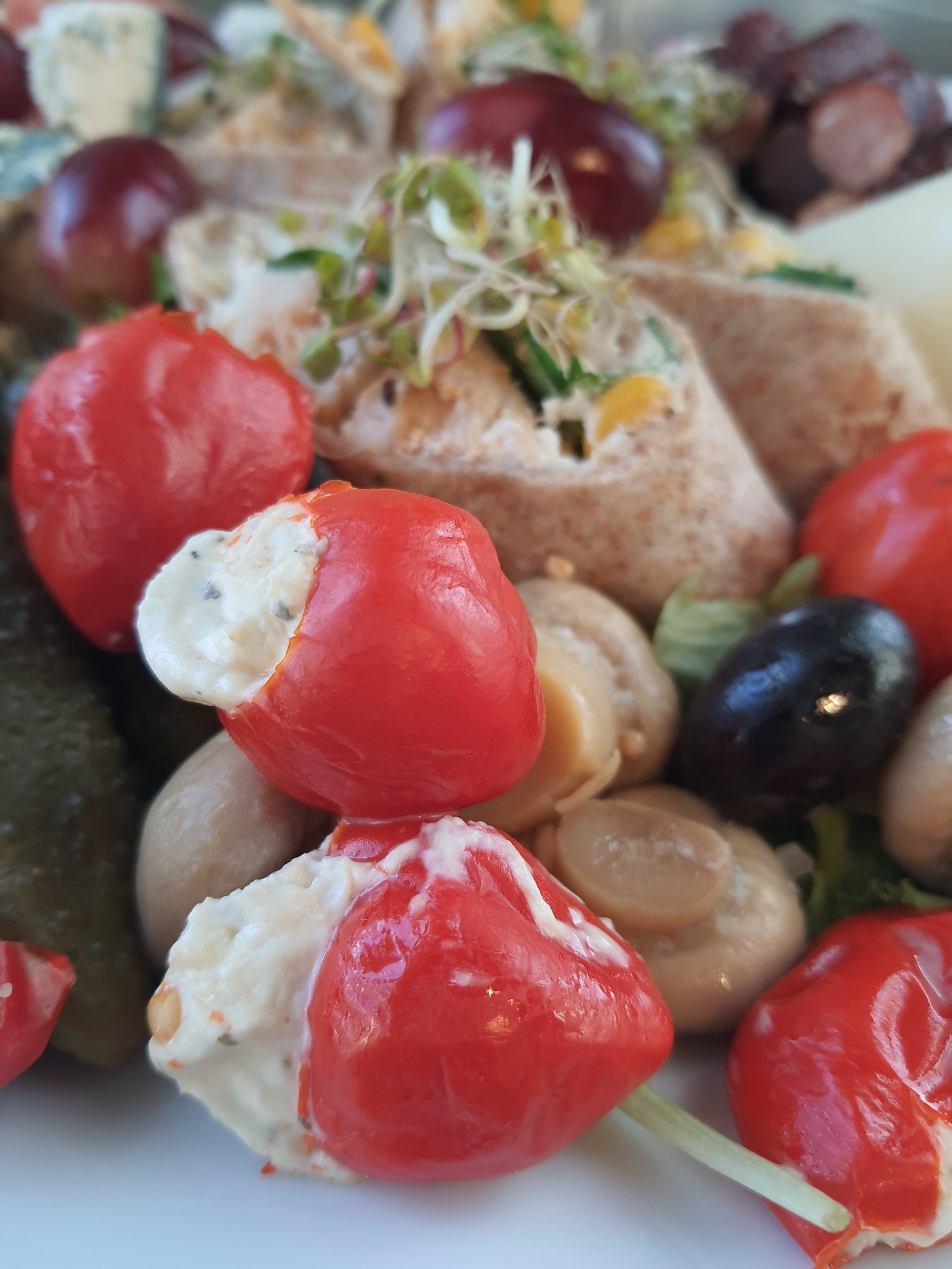 Dekoracyjna kompozycja cateringowa: czerwone papryczki nadziewane kremowym serem, mini tortille ze świeżymi kiełkami, marynowane pieczarki, ciemne winogrona i kawałki sera pleśniowego na białym...