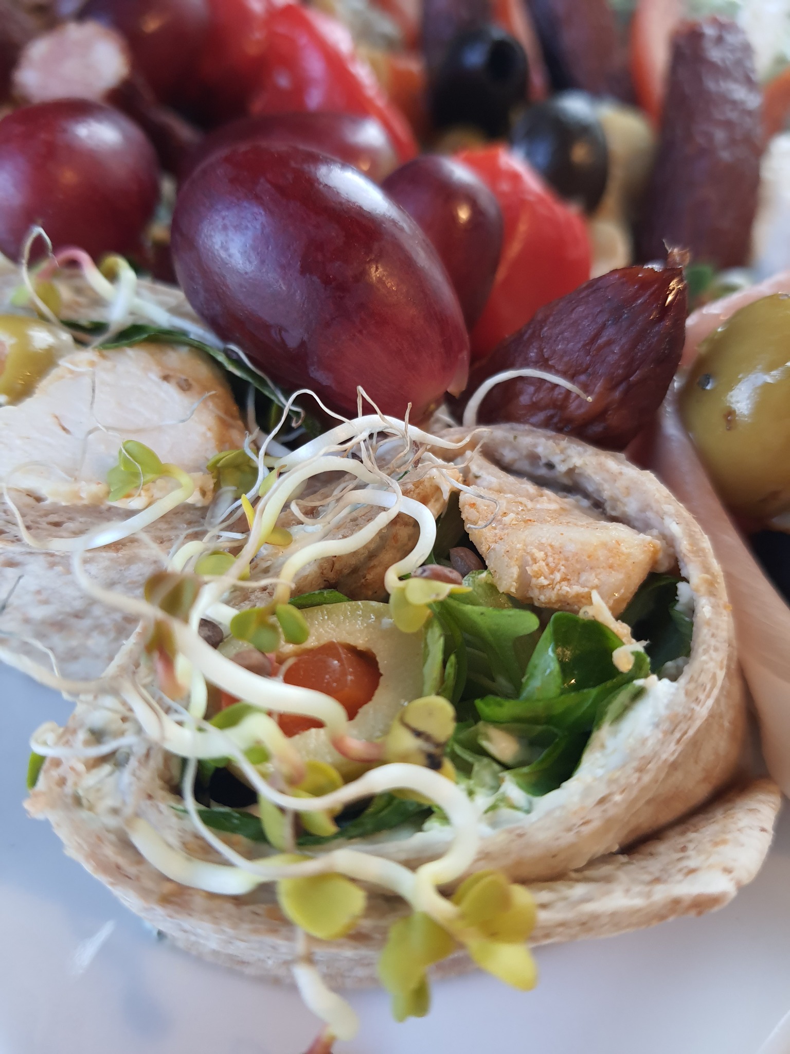 Zbliżenie na pitę wypełnioną kurczakiem, sałatą, pomidorkami koktajlowymi i kiełkami, z winogronami, oliwkami i salami w tle.