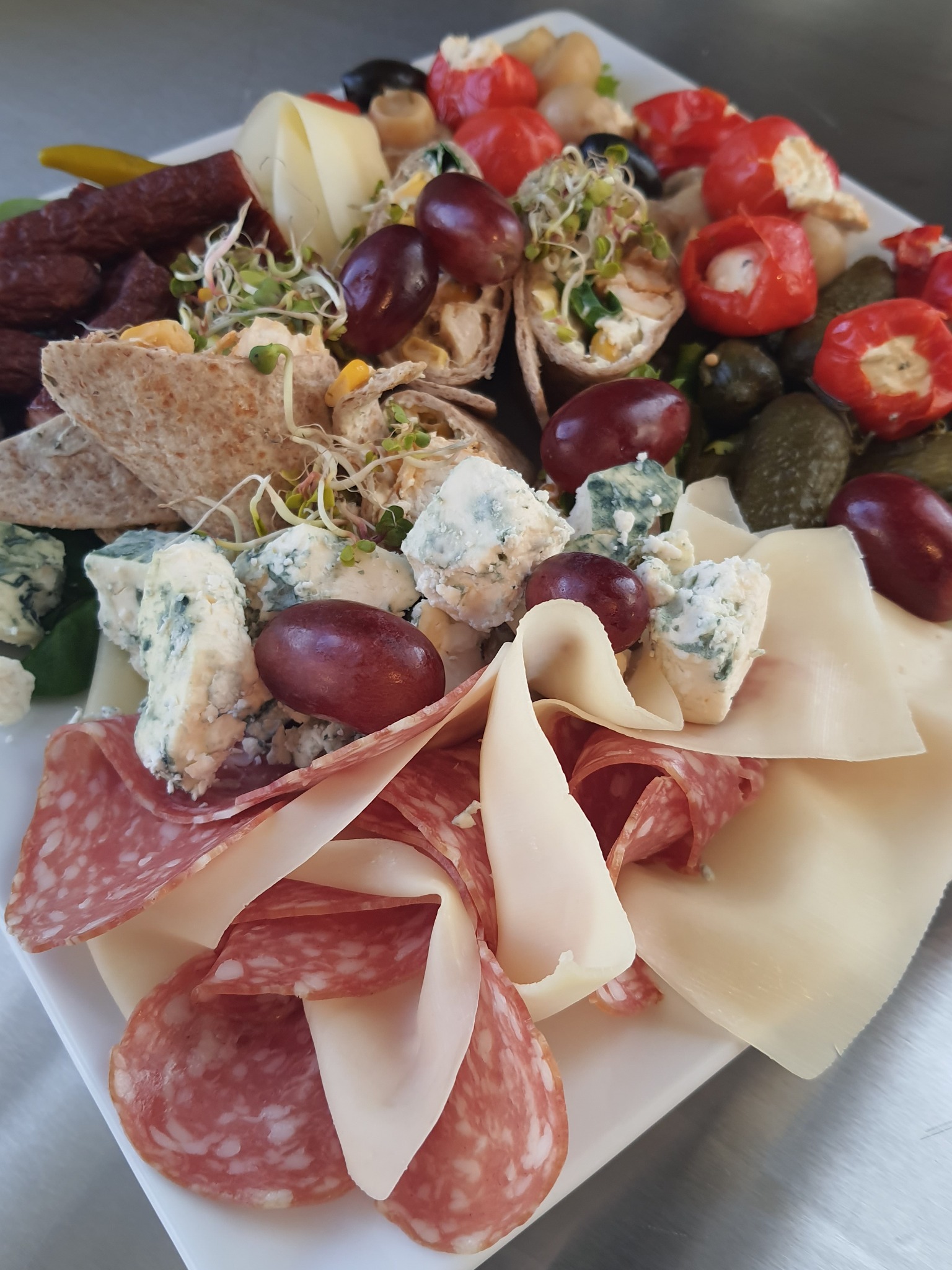 Deska serów i wędlin z dodatkami: salami, ser pleśniowy, winogrona, roladki z tortilli z kiełkami, papryczki faszerowane i korniszony na białym talerzu.