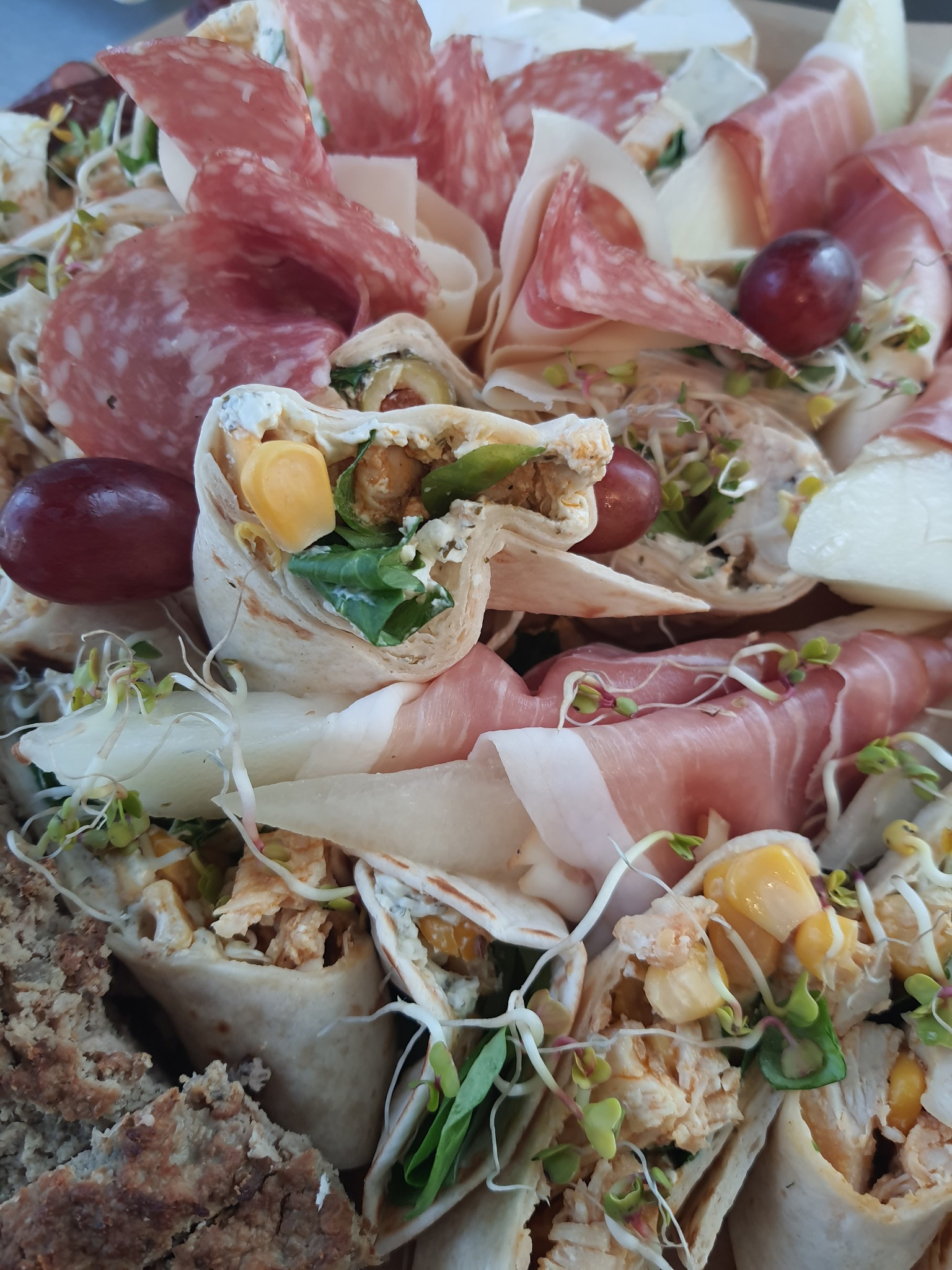 Dekoracyjna kompozycja przekąsek: roladki z tortilli z kurczakiem i kukurydzą, salami, szynka parmeńska, ser i winogrona, ozdobione świeżymi kiełkami.