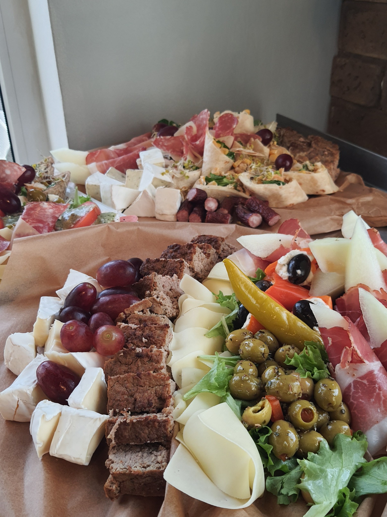 Bogato zaaranżowana deska serów, wędlin, pieczonego pasztetu i owoców, z widocznymi zielonymi oliwkami, papryką, melonem, winogronami i tortillą, ułożona na papierze kraft.