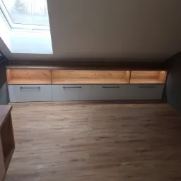 Szafki wnękowe z szarymi frontami i drewnianymi wnękami, oświetlone, zamontowane pod skosem dachu z oknem. Podłoga z paneli imitujących drewno.