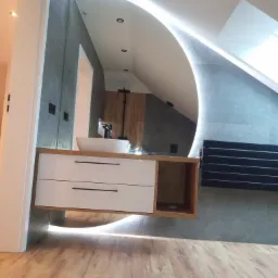 Nowoczesna łazienka na poddaszu: umywalka nablatowa na szafce z szufladami, podświetlane lustro owalne, drewniana podłoga, szare płytki.