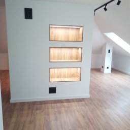 Expert wykończenia wnętrz - Wnętrze poddasza z drewnianą podłogą i wnękami ściennymi z oświetleniem LED. Minimalistyczny design, białe ściany, czarne akcenty oświetleniowe. Ukośne okno dachowe.
