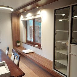Expert wykończenia wnętrz - Nowoczesne wnętrze: drewniany parapet-siedzisko z poduszką przy oknie, przeszklona szafa, drewniany stół z krzesłami. Ciepłe oświetlenie punktowe i minimalistyczny design.