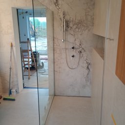 Expert wykończenia wnętrz - Nowoczesna kabina prysznicowa walk-in z marmurowym wykończeniem ścian, widok na zewnątrz przez otwarte drzwi. Minimalistyczny design i jasna kolorystyka.