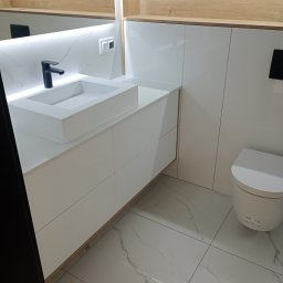 Expert wykończenia wnętrz - Nowoczesna łazienka z białą armaturą i szafkami pod umywalką, oświetlenie LED, płytki imitujące marmur na podłodze i ścianach, czarna bateria umywalkowa.