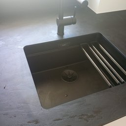 Expert wykończenia wnętrz - Czarny, nowoczesny zlew Blanco z baterią w blacie o szorstkiej fakturze. Minimalistyczny design, metalowe ociekacze. Górne światło podkreśla strukturę powierzchni blatu.