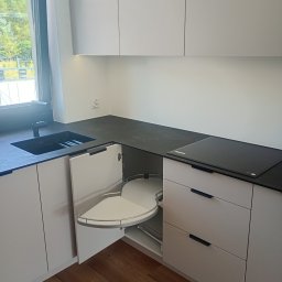 Expert wykończenia wnętrz - Biała, nowoczesna kuchnia z czarnym blatem i narożną szafką z wysuwanym systemem półek. Widoczny zlew i płyta indukcyjna. Jasne drewno na podłodze.