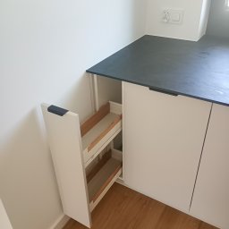 Expert wykończenia wnętrz - Biała szafka kuchenna z wysuwanym cargo, drewniane półki. Blat w odcieniach szarości, widoczna podłoga z paneli. Minimalistyczny design, nowoczesne wykończenie.