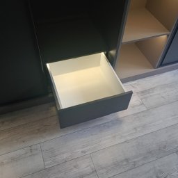 Expert wykończenia wnętrz - Szara szafa wnękowa z wysuniętą szufladą, widok z bliska na podłogę w panelach w Tarnowie. Oświetlenie wnętrza szafy dodaje ciepła.