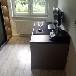 Expert wykończenia wnętrz - Nowoczesny pokój z biurkiem, komputerem i szafą wnękową. Minimalistyczny design, jasne panele podłogowe i duże okno z widokiem na zieleń. Szafa z oświetleniem.