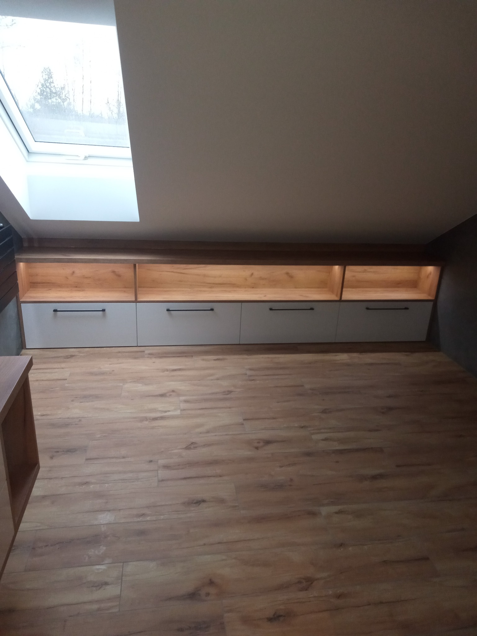 Szafki wnękowe z szarymi frontami i drewnianymi wnękami, oświetlone, zamontowane pod skosem dachu z oknem. Podłoga z paneli imitujących drewno.