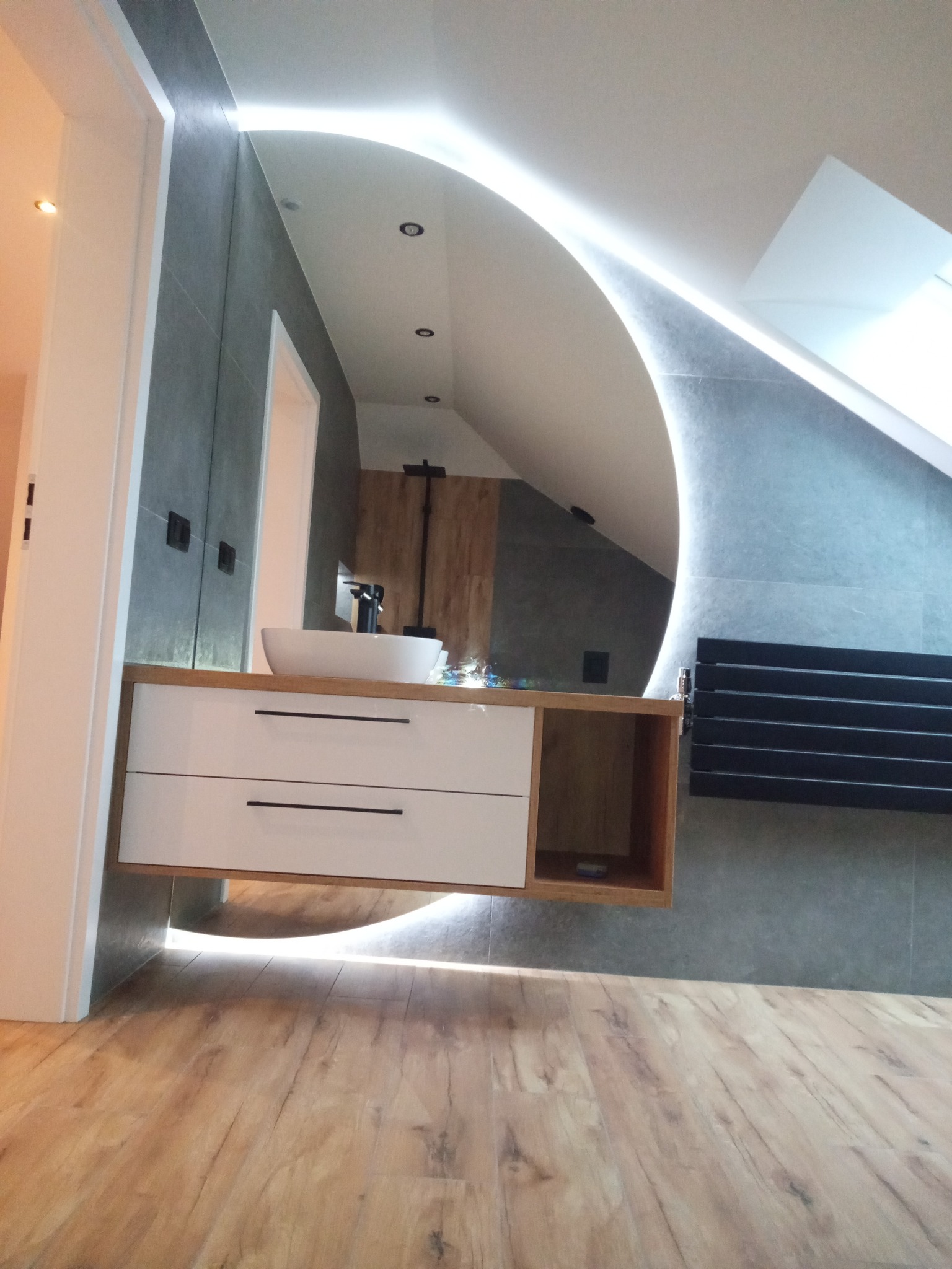 Nowoczesna łazienka na poddaszu: umywalka nablatowa na szafce z szufladami, podświetlane lustro owalne, drewniana podłoga, szare płytki.