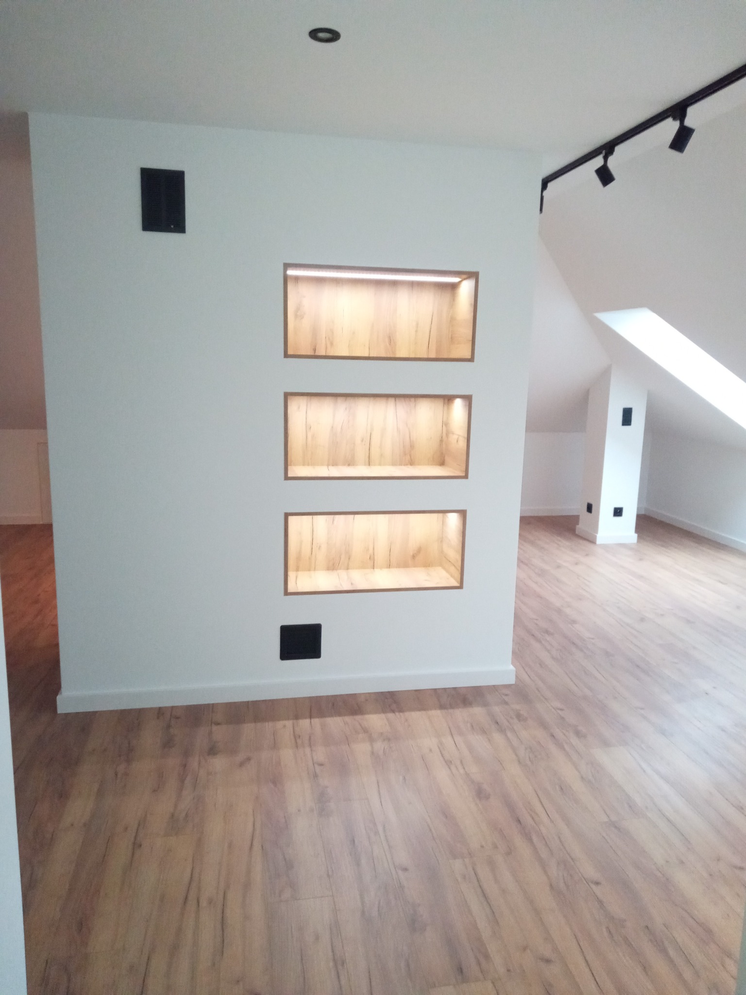 Wnętrze poddasza z drewnianą podłogą i wnękami ściennymi z oświetleniem LED. Minimalistyczny design, białe ściany, czarne akcenty oświetleniowe. Ukośne okno dachowe.