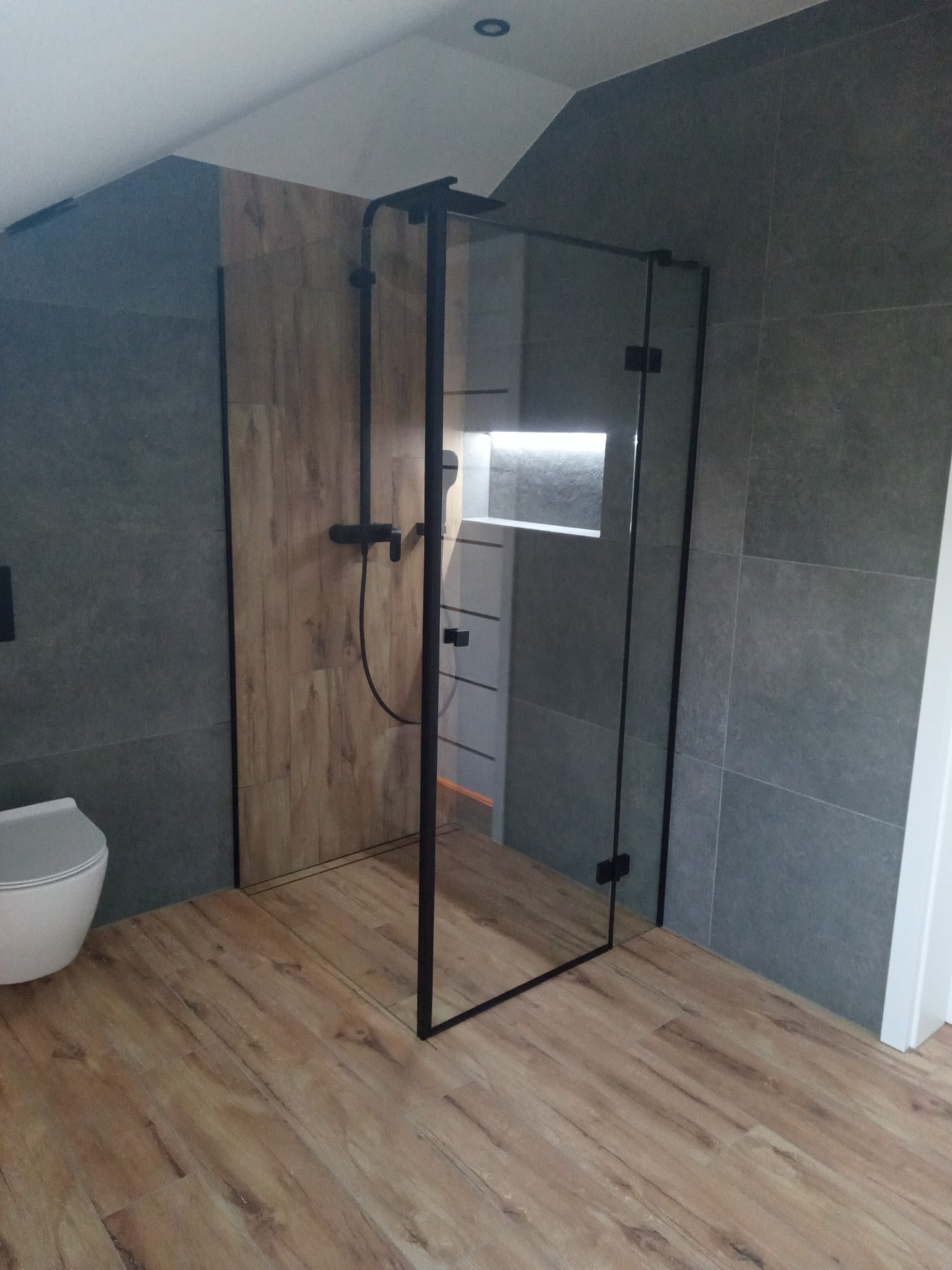 Nowoczesna łazienka na poddaszu z kabiną prysznicową typu walk-in, wykończona szarą płytką i drewnopodobną okładziną. Widoczna toaleta i drewniana podłoga.