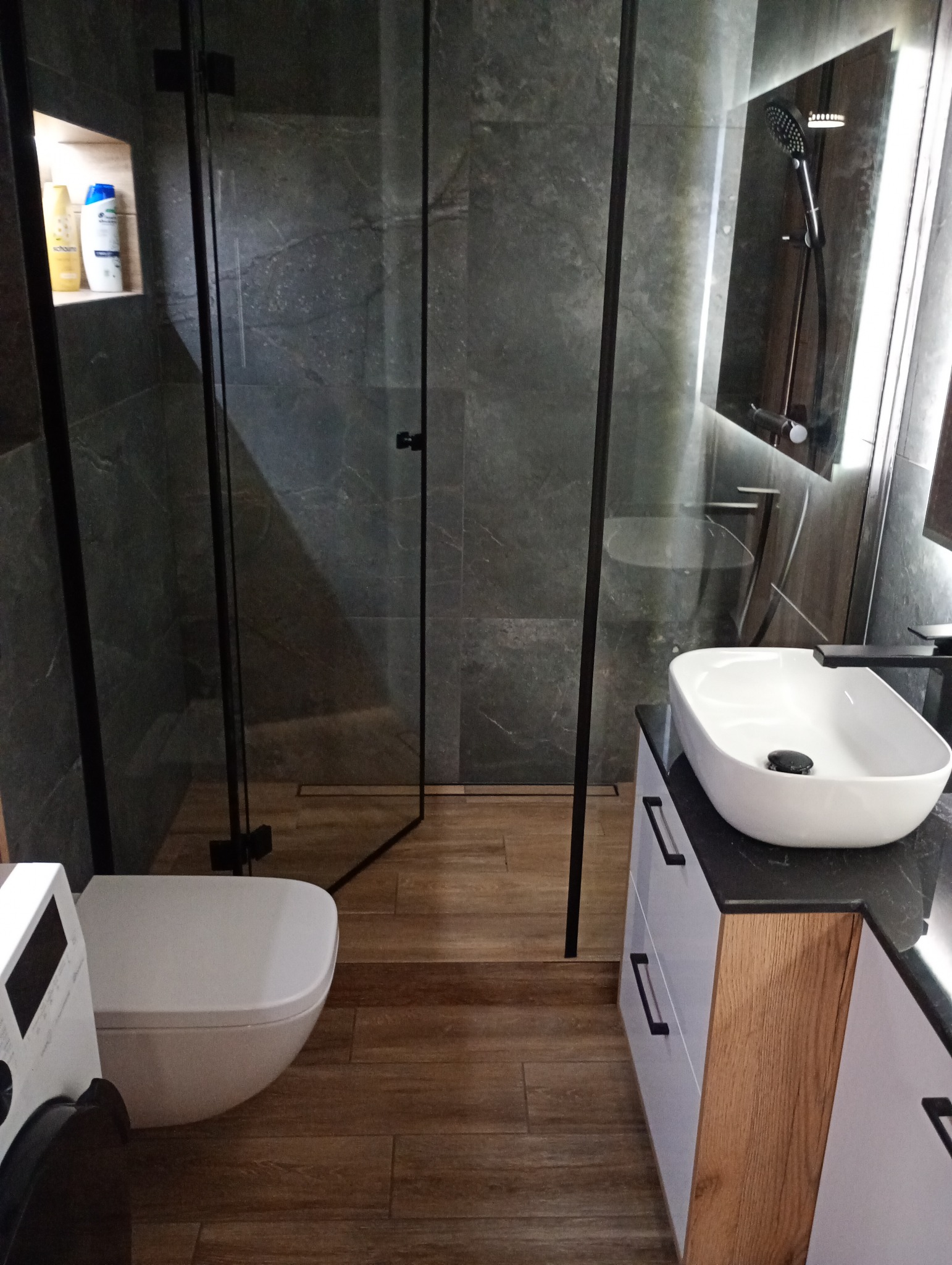 Nowoczesna łazienka z kabiną prysznicową typu walk-in, umywalką nablatową na szafce i toaletą podwieszaną. W tle szare płytki imitujące kamień i drewnopodobna podłoga.