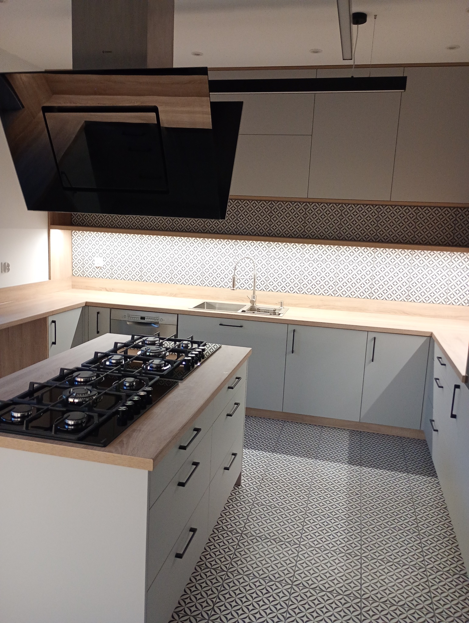 Nowoczesna kuchnia w kształcie litery U z wyspą i czarną płytą gazową. Jasne szafki, wzorzyste płytki nad blatem i na podłodze. Czarny okap nad płytą.