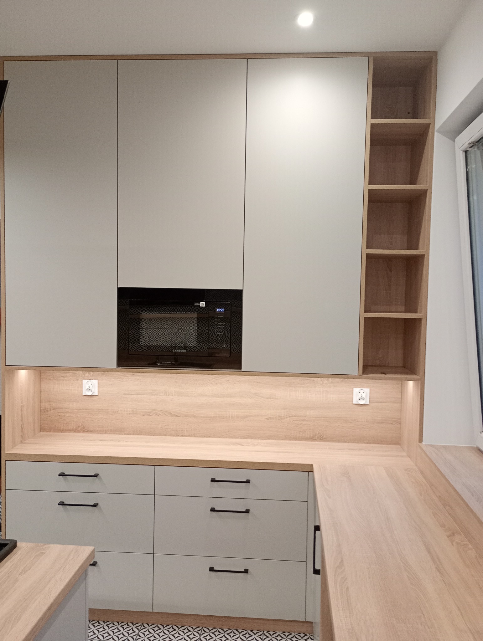 Jasnoszara zabudowa kuchenna z drewnianym blatem i wbudowaną mikrofalówką. Na podłodze wzorzyste płytki. Oświetlenie LED pod szafkami.