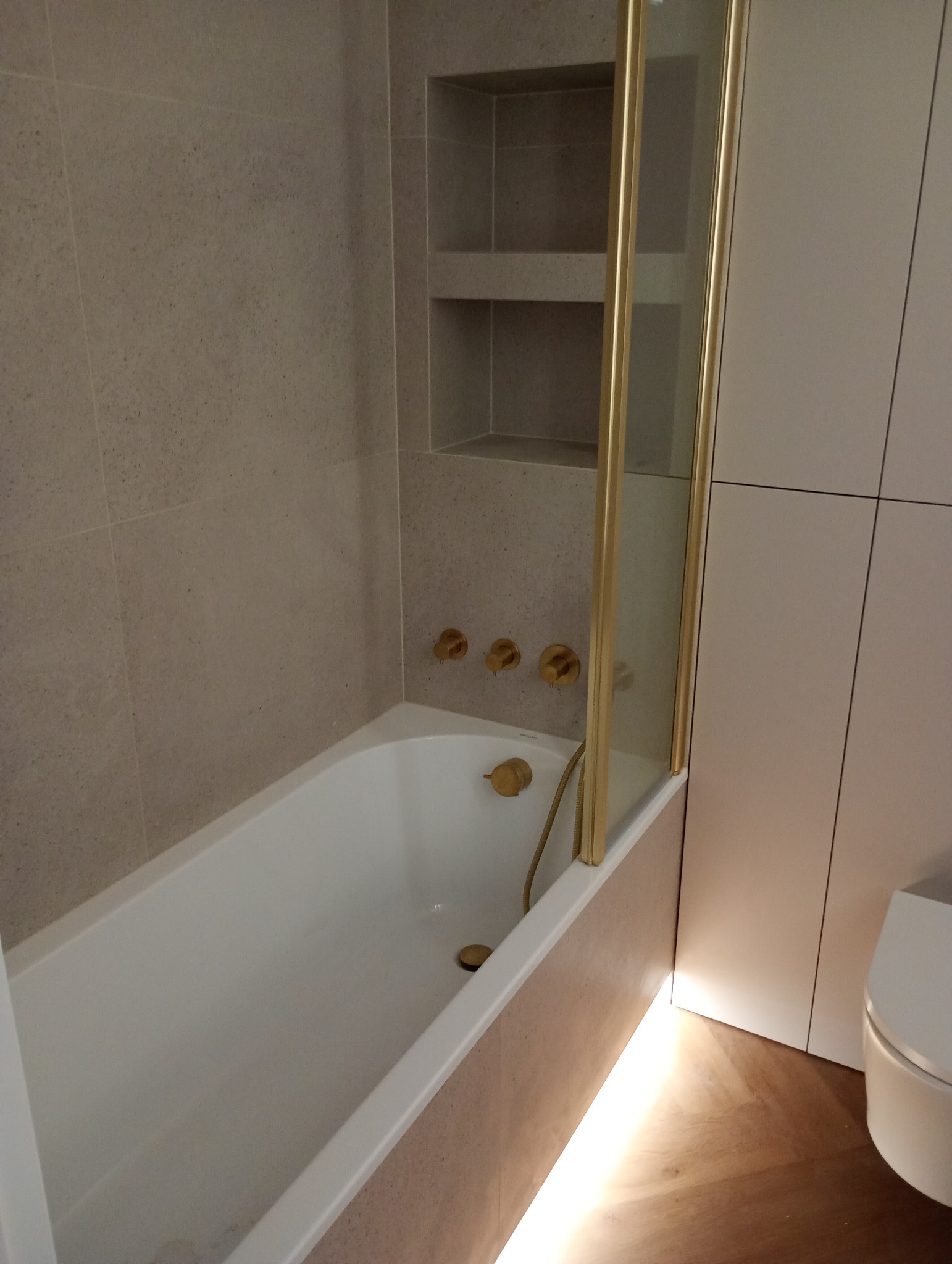 Wykończona łazienka: biała wanna z złotą armaturą, szklany parawan nawannowy i wnęka w ścianie z półkami, podświetlenie LED przy podłodze. Minimalistyczny design.