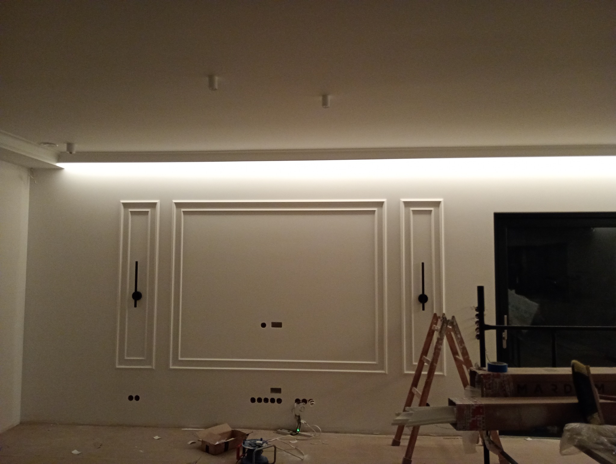 Ściana z dekoracyjnymi listwami i podświetleniem LED w trakcie montażu, widoczne gniazdka elektryczne i drabina. Wnętrze w trakcie wykańczania.