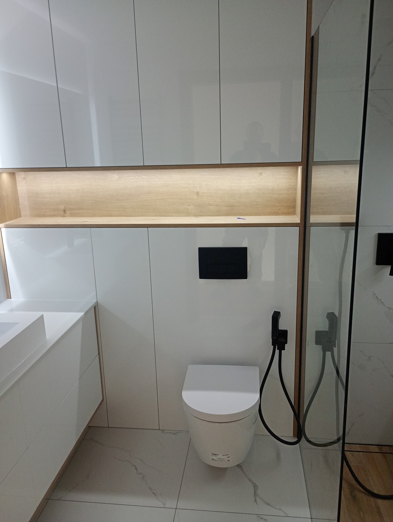 Nowoczesna łazienka z białymi, lakierowanymi szafkami, podświetlaną półką i toaletą z bidetką. Marmurowe płytki podłogowe. Minimalistyczny design.