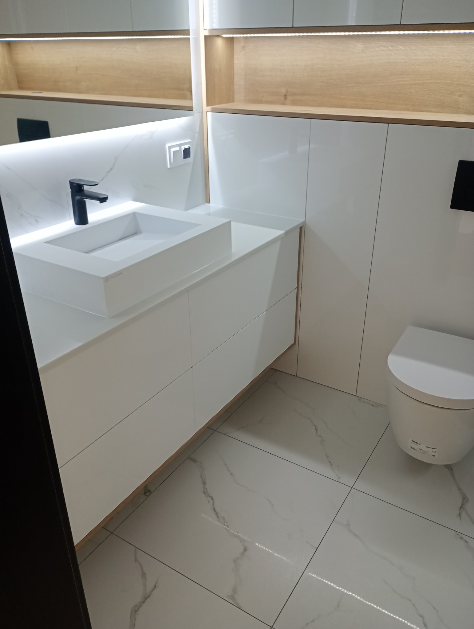 Nowoczesna łazienka z białą armaturą i szafkami pod umywalką, oświetlenie LED, płytki imitujące marmur na podłodze i ścianach, czarna bateria umywalkowa.
