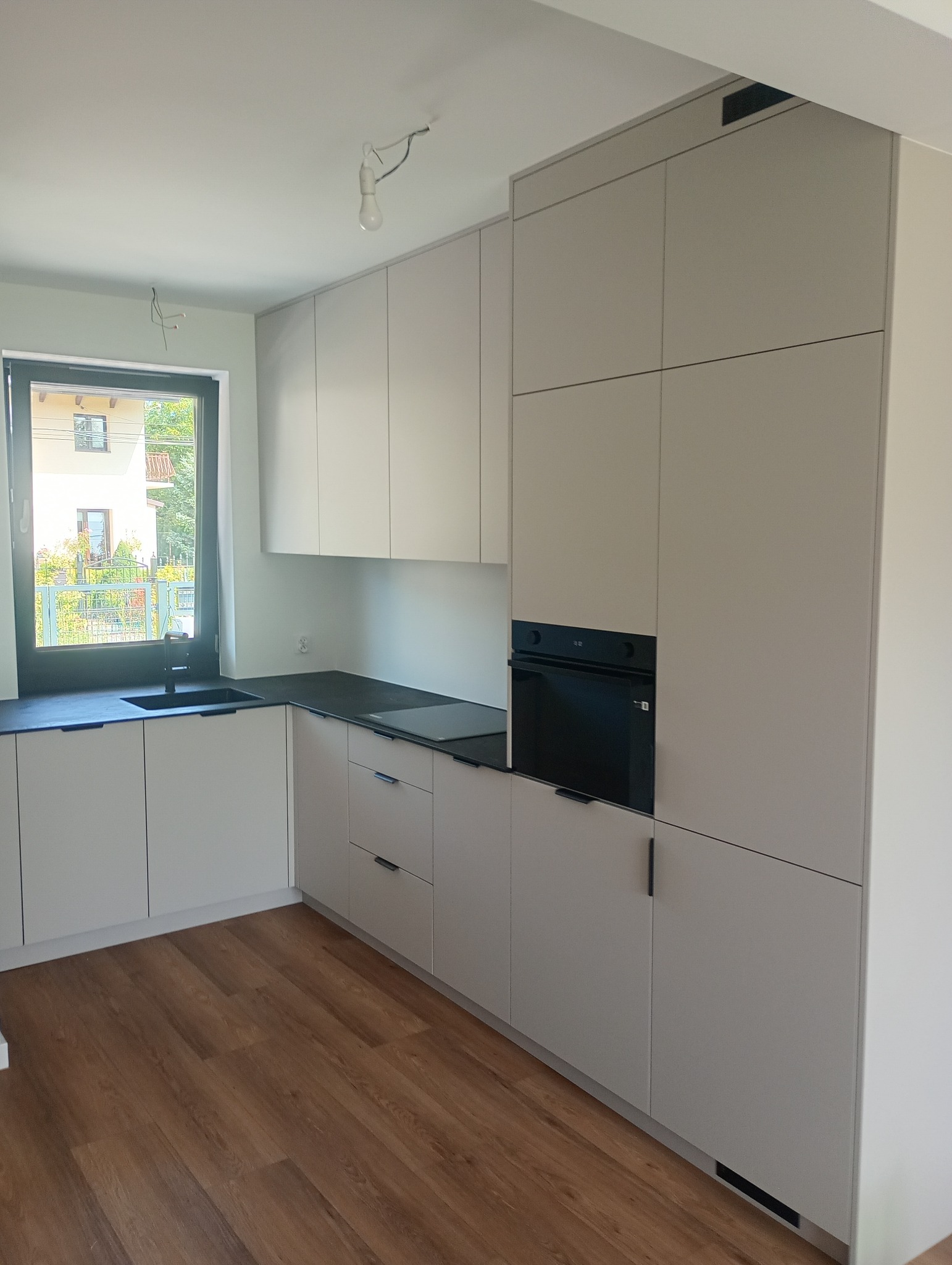 Jasna kuchnia z zabudową pod sufit, czarnym blatem i drewnianą podłogą. Widok na zewnątrz przez okno z czarną ramą. Minimalistyczny design.