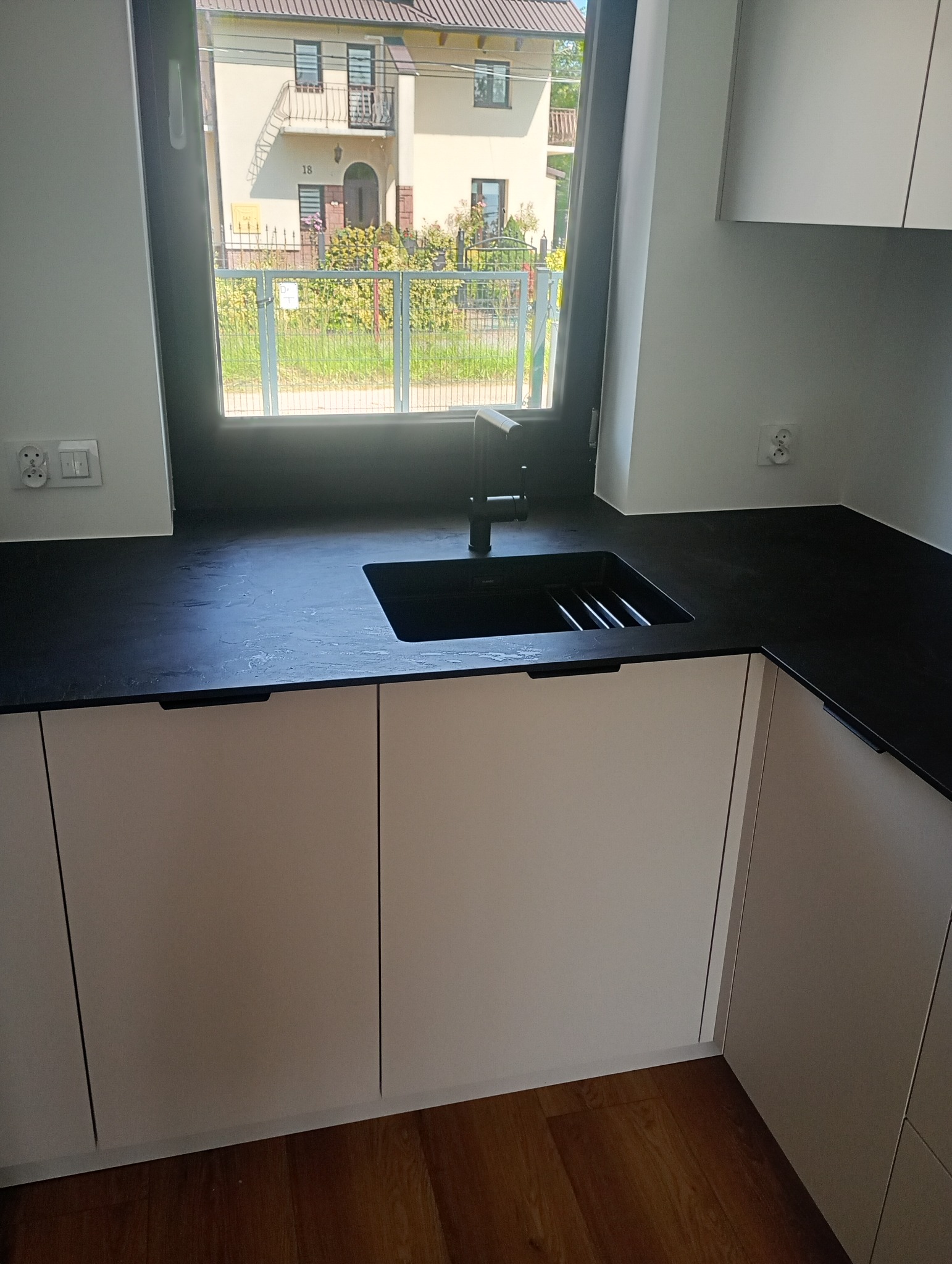 Minimalistyczna kuchnia: białe szafki z czarnym blatem i zlewem pod oknem z widokiem na dom. Nowoczesny design, proste linie, jasne wnętrze.