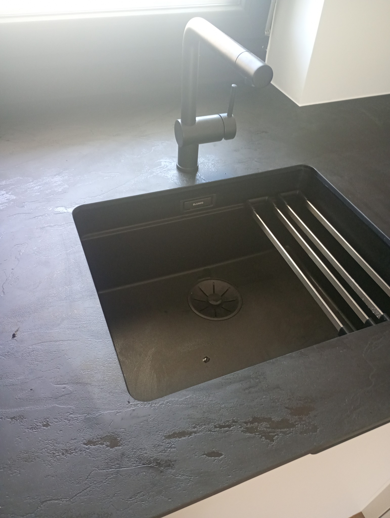 Czarny, nowoczesny zlew Blanco z baterią w blacie o szorstkiej fakturze. Minimalistyczny design, metalowe ociekacze. Górne światło podkreśla strukturę powierzchni blatu.