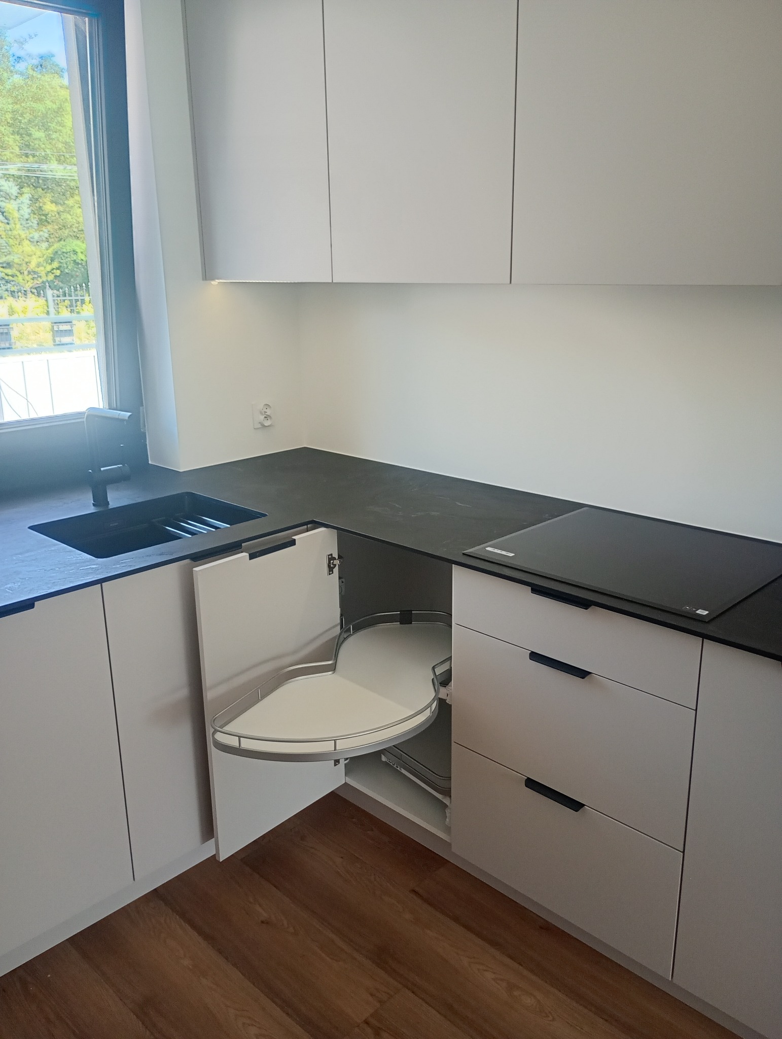 Biała, nowoczesna kuchnia z czarnym blatem i narożną szafką z wysuwanym systemem półek. Widoczny zlew i płyta indukcyjna. Jasne drewno na podłodze.