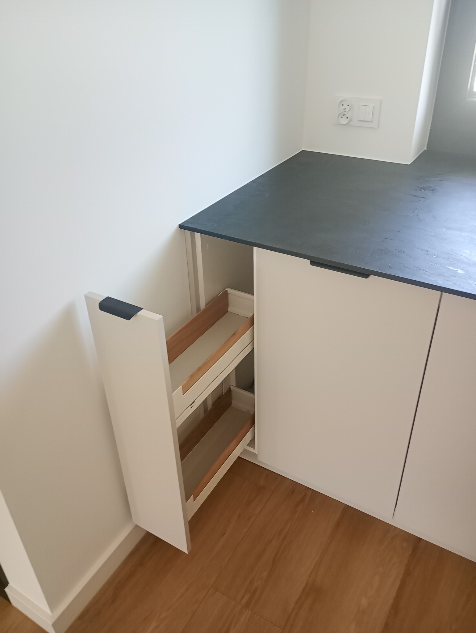 Biała szafka kuchenna z wysuwanym cargo, drewniane półki. Blat w odcieniach szarości, widoczna podłoga z paneli. Minimalistyczny design, nowoczesne wykończenie.