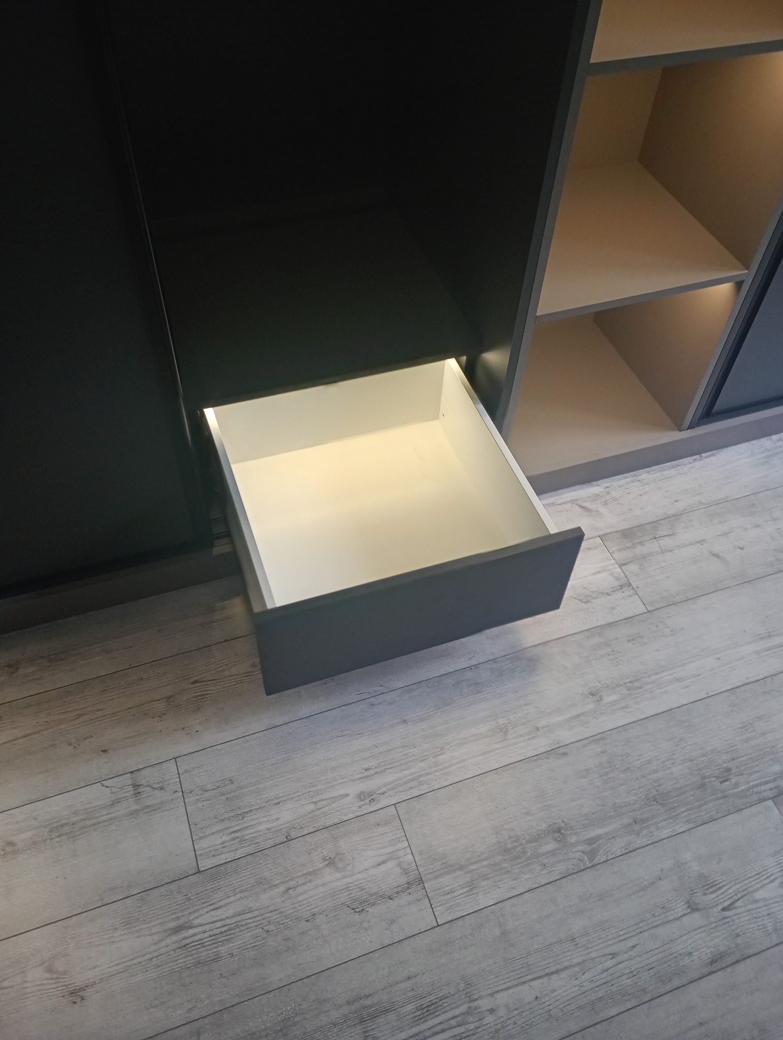 Szara szafa wnękowa z wysuniętą szufladą, widok z bliska na podłogę w panelach w Tarnowie. Oświetlenie wnętrza szafy dodaje ciepła.